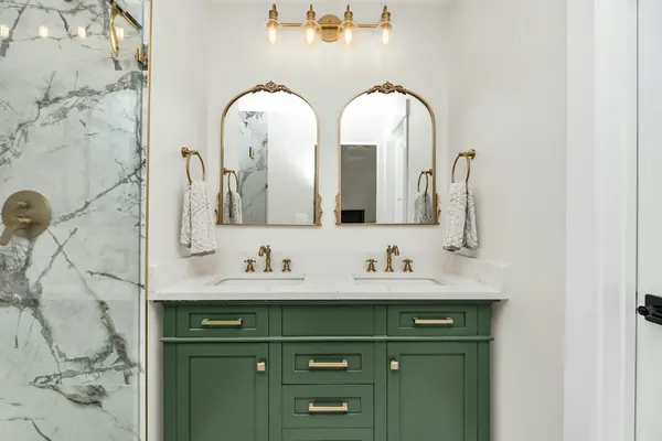 Green double vanity with brass faucets beside a marble-look shower wall