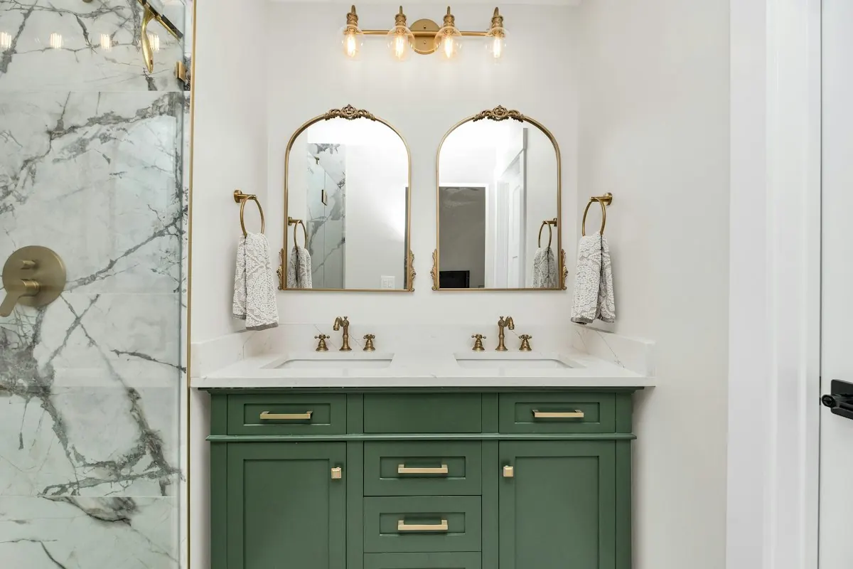 Green double vanity with brass faucets and arched mirrors beside a marble-look shower wall