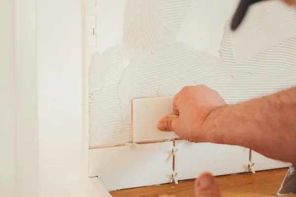 Hands pressing white wall tiles into place with spacers above a wood countertop