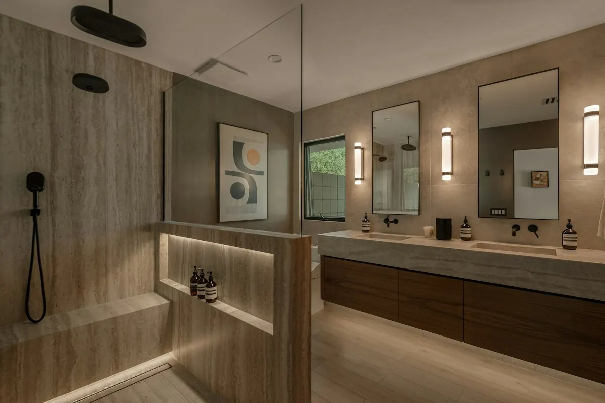 Large walk-in shower with a lit bench niche behind a half wall, beside a double vanity